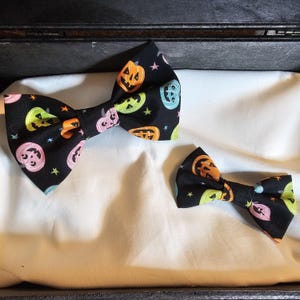May include: Two bow ties made of black fabric featuring a repeating pattern of jack-o'-lanterns in shades of orange, pink, green, and blue, interspersed with small colorful stars. The bow ties are presented in two different sizes side by side.