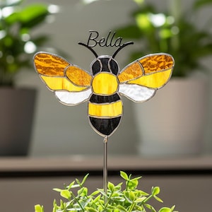May include: A decorative bee garden stake with the name "Bella" on top. The bee has yellow, white, and black stained glass wings and body. It is placed in a white pot with a green plant. The image is taken indoors.