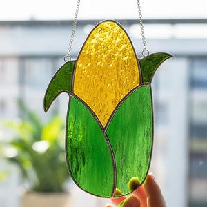 Könnte beinhalten: Ein Glasfenster-Dekoration in Form eines Maiskolbens mit gelben Körnern und grünen Blättern, an einer silbernen Kette aufgehängt. Der Mais wird von einer Hand vor einem Fenster mit Pflanzen gehalten. Der Text "Blselling" befindet sich unten rechts.