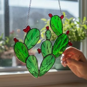 May include: A stained glass cactus with green leaves and red accents hangs from a silver chain. The cactus is suspended in front of a window, casting a shadow on a wooden surface. The cactus is approximately 6 inches tall.