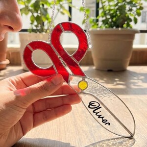 May include: A stained glass scissor-shaped suncatcher with red handles and a clear blade. The name "Oliver" is written on the blade. The suncatcher hangs from a silver chain. The logo "Blselling" is visible in the bottom right corner.