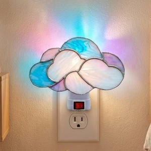 May include: A cloud-shaped night light with stained glass panels in shades of blue, white, and purple is plugged into a wall outlet. The light is on, casting a soft glow. A sleeping corgi puppy rests on a fluffy rug near the outlet.