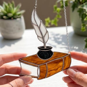 May include: Stained glass book with a quill and inkwell design. The book is brown with a clear glass page, and the inkwell is black. A white feather quill sits above the inkwell. The piece is suspended by a silver chain. The overall theme is literary.