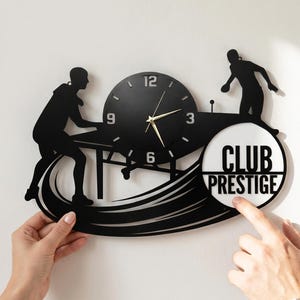 Puede incluir: Reloj de pared de metal negro con siluetas de jugadores de ping pong y las palabras "CLUB PRESTIGE". El reloj tiene una esfera redonda con manecillas doradas y un marco negro. El reloj está sostenido por dos manos.