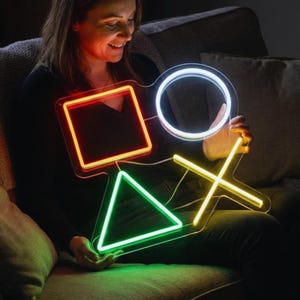 May include: A neon sign featuring the PlayStation controller symbols: a red square, a white circle, a green triangle, and a yellow X. The sign is illuminated and held by a person sitting on a couch. The sign is a vibrant and colorful decorative item.