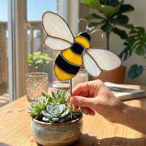 Pode incluir: Uma estaca de jardim decorativa de vitral com uma abelha com listras amarelas e pretas e asas brancas. A abelha é colocada em um pequeno vaso de cerâmica com suculentas.
