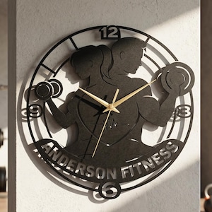 May include: Black metal wall clock with a fitness theme. The clock features silhouettes of two people lifting weights, with the words "Anderson Fitness" and the numbers 12, 3, 6, and 9. The clock hands are gold.