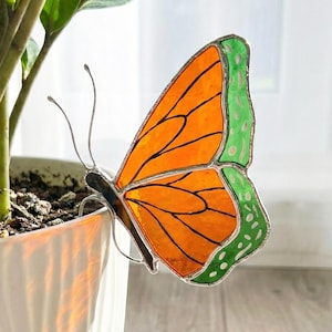 May include: A stained glass monarch butterfly decoration with orange and green wings. The butterfly is attached to a white ceramic pot with a green plant. The text "Monarch Butterfly 1" is visible in the upper right corner.