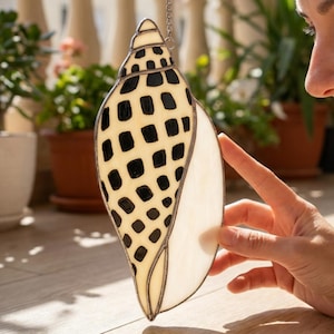 May include: Stained glass seashell suncatcher with a cream and black spotted pattern. The decorative shell is hanging from a silver chain. The shell is a decorative item for the home.