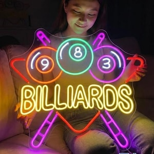 May include: A neon sign with the word "BILLIARDS" in yellow, and three billiard balls with the numbers 3, 8, and 9. The sign has a red, pink, and purple outline. The sign is held by a person.