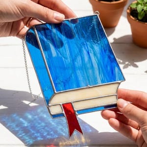 May include: A stained glass book-shaped suncatcher with a blue and white design. The book has a red ribbon bookmark and a silver chain for hanging. The suncatcher is held by two hands, with sunlight casting a blue shadow.