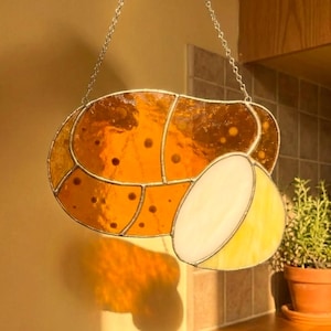 May include: Stained glass potato-shaped suncatcher with a chain for hanging. The suncatcher is made of amber and yellow glass, with a section cut out to reveal a lighter yellow interior. The design includes dark spots.