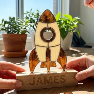 May include: A stained glass rocket ship in cream and brown, set on a wooden base engraved with the name "JAMES". The rocket features a circular window and detailed fins. The rectangular base is made of light-coloured wood. The overall design is decorative.