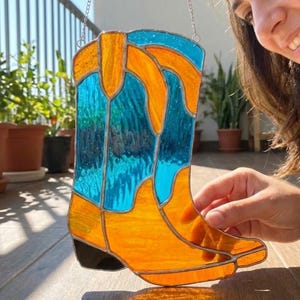 May include: Stained glass cowboy boot in orange and turquoise blue. The decorative boot has a black heel and is suspended by a silver chain. The boot is made of glass and is designed to be hung as a decorative item.