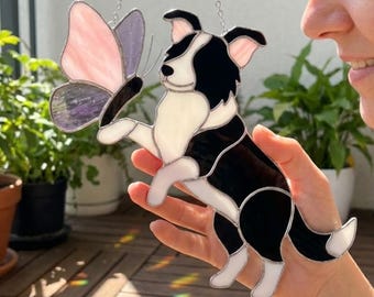 May include: A stained glass decoration featuring a black and white dog interacting with a pink and purple butterfly. The dog is in a playful pose, with its paw extended towards the butterfly. The piece is suspended by a silver chain.