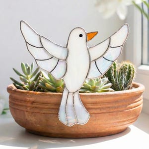 May include: A stained glass dove with outstretched wings, featuring white and iridescent glass panels, and an orange beak. The dove is set in a terracotta pot filled with succulents and a cactus. The word "Dove" is in the upper left corner.