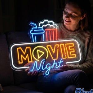 May include: A neon sign with the words "MOVIE Night" in orange and blue, with a play button symbol. The sign also features neon images of popcorn and a drink. The sign is held by a person.