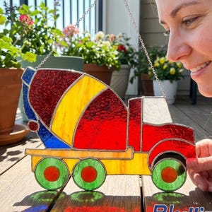 Op de afbeelding: Een kleurrijke glas-in-lood betonmixer met rode, gele en blauwe secties. De vrachtwagen heeft groene wielen met rode centra en wordt opgehangen aan een zilveren ketting. De achtergrond toont potplanten en een houten terras.