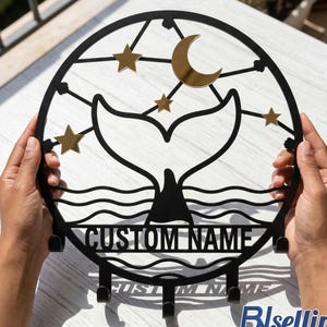 May include: Black metal wall art featuring a whale tail design with a crescent moon and stars. The art piece includes hooks for hanging items and the text "CUSTOM NAME". The design is held by two hands.