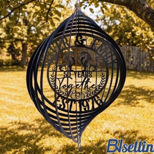 May include: Black metal wind spinner with a decorative design. The spinner features the text "Forever & Family" and "50 Years Together EST 1976" with two lion figures. The spinner is hanging from a tree branch.