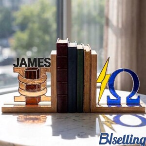 May include: A set of wooden bookends with a stack of books. One bookend features the name "JAMES" in black letters. The other bookend has a yellow lightning bolt and a blue Omega symbol. The bookends are on a white surface.