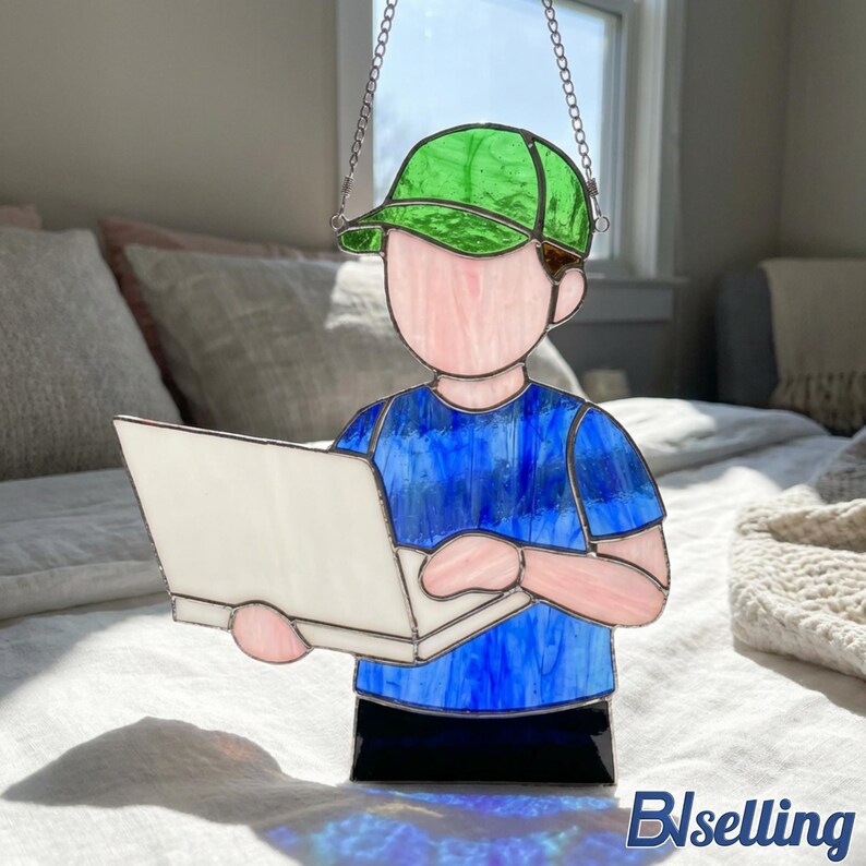 May include: Stained glass figure of a person wearing a green cap and blue shirt, holding a white laptop. The figure is suspended by a chain. The figure's face and arms are pink, and the base is black. The image is set against a soft background.