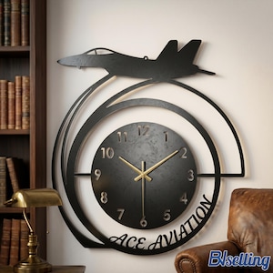 Op de afbeelding: Zwarte metalen wandklok met een straaljager-silhouet boven een cirkelvormige wijzerplaat. De klok heeft gouden wijzers en de woorden "ACE AVIATION" onderaan. De klok is aan een muur bevestigd.