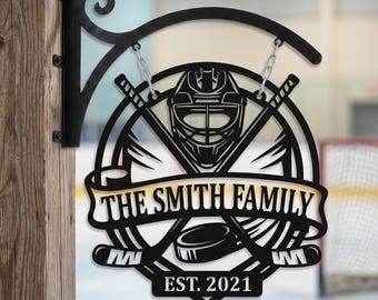 Puede incluir: Un letrero circular de metal negro con una máscara de portero de hockey, dos palos de hockey cruzados y un disco. Una pancarta en el centro muestra el texto THE SMITH FAMILY, con EST. 2021 debajo. El letrero cuelga de un soporte de metal.