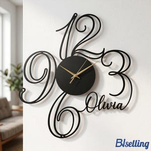 May include: Black metal wall clock with gold hands and the name "Olivia" in cursive script. The clock features large, stylized numbers 12, 3, 6, and 9. The clock is mounted on a white wall.
