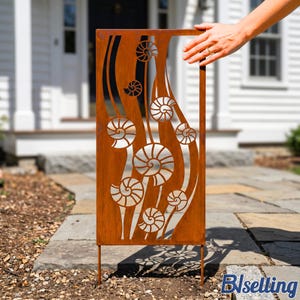 May include: A rectangular, weathered-look metal garden screen with a cut-out design of stylized nautilus shells and flowing lines. The screen is brown with a rust-like finish and has two metal stakes for ground placement. A hand is touching the top frame.