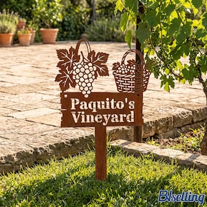Può includere: Un cartello da giardino in metallo color ruggine con un design ritagliato di un grappolo d'uva con foglie e un cesto di uva. Il cartello rettangolare mostra il testo bianco Paquito's Vineyard ed è sostenuto da un picchetto in metallo.