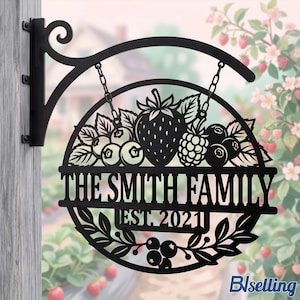 May include: A black metal circular hanging sign featuring a cutout design of strawberries, blueberries, and raspberries. The text reads THE SMITH FAMILY and EST. 2021. The sign is suspended by two chains from a decorative wall-mounted bracket.