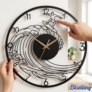 May include: Black metal wall clock featuring a surfer riding a wave. The clock has a black face with gold hands and a black circular frame. The numbers are in a simple, modern font. The clock is being held up against a white wall.