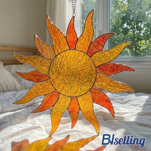 May include: A sun-shaped stained glass suncatcher featuring a textured yellow center surrounded by alternating yellow and orange pointed rays. The piece is constructed with dark metal lead lines and hangs from a silver-colored metal chain.