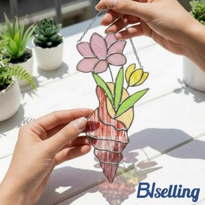 May include: A stained glass art piece featuring a pink lotus flower, yellow tulip, and green leaves. The design is set within a red and white striped seashell shape, suspended by a silver chain. The artwork is held up by hands.