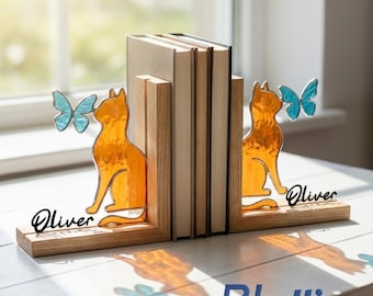 Puede incluir: Un par de sujetalibros de madera con siluetas de gatos de vidrio tintado naranja y mariposas de vidrio tintado azul claro. Cada base de madera tiene grabado el nombre Oliver en letra cursiva negra, sosteniendo dos libros.