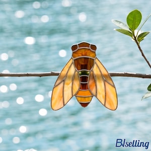 May include: A stained glass cicada ornament featuring amber, orange, and brown glass segments held together by thin metal solder lines. The insect-shaped piece is perched on a thin horizontal twig, showcasing translucent wings and a segmented body.