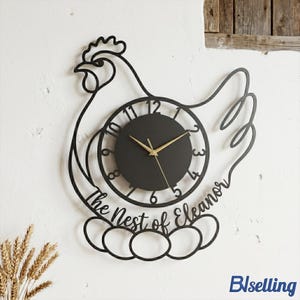 May include: Black metal wall clock in the shape of a hen, with a round clock face and gold hands. The clock features the text "The Nest of Eleanor" and is designed with a hen and egg motif.