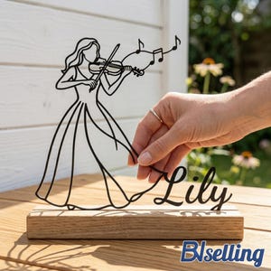 May include: Black metal sculpture of a woman playing a violin, mounted on a wooden base. The sculpture includes musical notes and the name "Lily" in cursive. The base also has the text "Blselling".