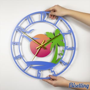 May include: A blue, laser-cut wall clock with a beach theme. The clock features a sunset, palm tree, and seagull design. The clock hands are gold. The clock is being held by two hands.