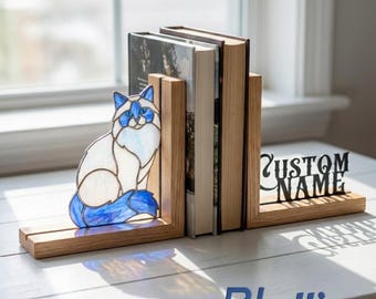 Puede incluir: Un juego de sujetalibros de madera con una figura de gato de vidrieras en tonos blancos y azules en un lado y un recorte de metal negro con el texto CUSTOM NAME en el otro, sosteniendo dos libros en una superficie clara.