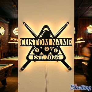 May include: A black metal wall sign featuring a backlit design of two crossed pool cues and a billiard ball rack. The sign displays the text "CUSTOM NAME" and "EST. 2026" with three billiard balls numbered 3, 8, and 1 integrated into the rack.