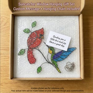 May include: A stained glass suncatcher gift set featuring a red cardinal and a blue hummingbird on a branch. The set includes a custom message and hanging chain. The gift set is packaged in a cardboard box.