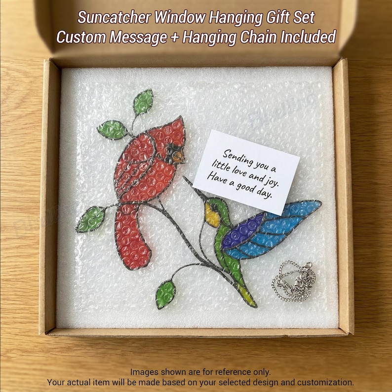 May include: A stained glass suncatcher gift set featuring a red cardinal and a blue hummingbird on a branch. The set includes a custom message and hanging chain. The gift set is packaged in a cardboard box.
