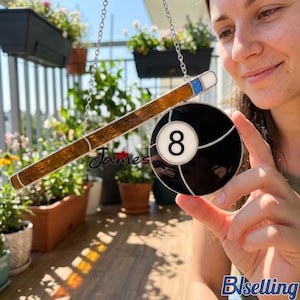 Könnte beinhalten: Ein Buntglas-Suncatcher mit Billard-Motiv, der einen braunen Billardqueue und eine schwarze Acht-Ball mit einer weißen 8 zeigt. Der Suncatcher wird von einer silbernen Kette gehalten. Das Wort "James" steht auf dem Queue.