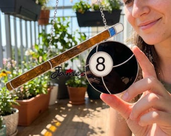 May include: A stained glass billiard-themed suncatcher featuring a brown cue stick and a black eight ball with a white number 8. The suncatcher is suspended by a silver chain. The word "James" is written on the cue stick.