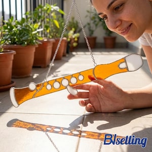 Puede incluir: Un atrapasoles de flauta de vidriera con un cuerpo naranja brillante y detalles blancos, suspendido por una cadena plateada. El atrapasoles proyecta una sombra sobre una superficie embaldosada, con plantas en macetas en el fondo.