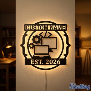 May include: A circular black metal wall sign with integrated backlighting. The design features a laptop, a computer monitor, two gears, and code brackets. The text CUSTOM NAME is at the top, and EST. 2026 is at the bottom. A white power cord hangs below.