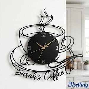 Può includere: Orologio da parete nero a forma di tazza da caffè, piattino e cucchiaio. Il quadrante dell'orologio è nero con lancette e numeri dorati. Le parole "Sarah's Coffee Co." sono scritte in corsivo.