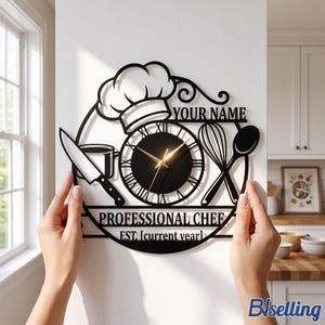 May include: A black metal wall clock featuring a chef hat, a knife, a whisk, and a spoon. The clock face has Roman numerals and gold hands. Text includes "YOUR NAME", "PROFESSIONAL CHEF", and "EST. [current year]". The design is circular and open-work.
